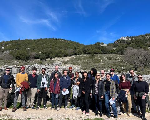 18 individuals pose for a group picture on a sunny day in front of ancient walls and a hilltop.