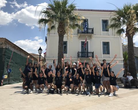 A group of 25 young people standing in front of a white building, all wearing the same T-shirts, with their hands raised and smiling.