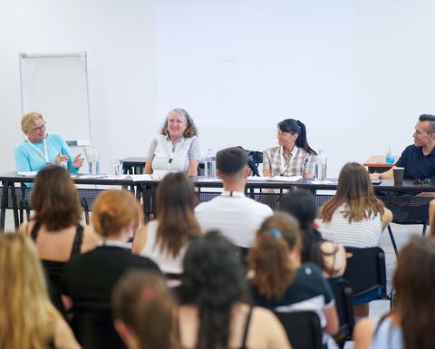 A panel of three women and one man in a classroom setting. They appear to engage in a discussion, and their audience listens attentively.
