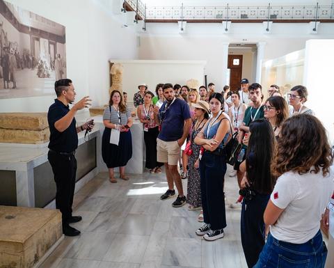 A group of people of various ages stands in a museum setting, listening attentively to Professor Stocking, who is speaking and gesturing with his hands.
